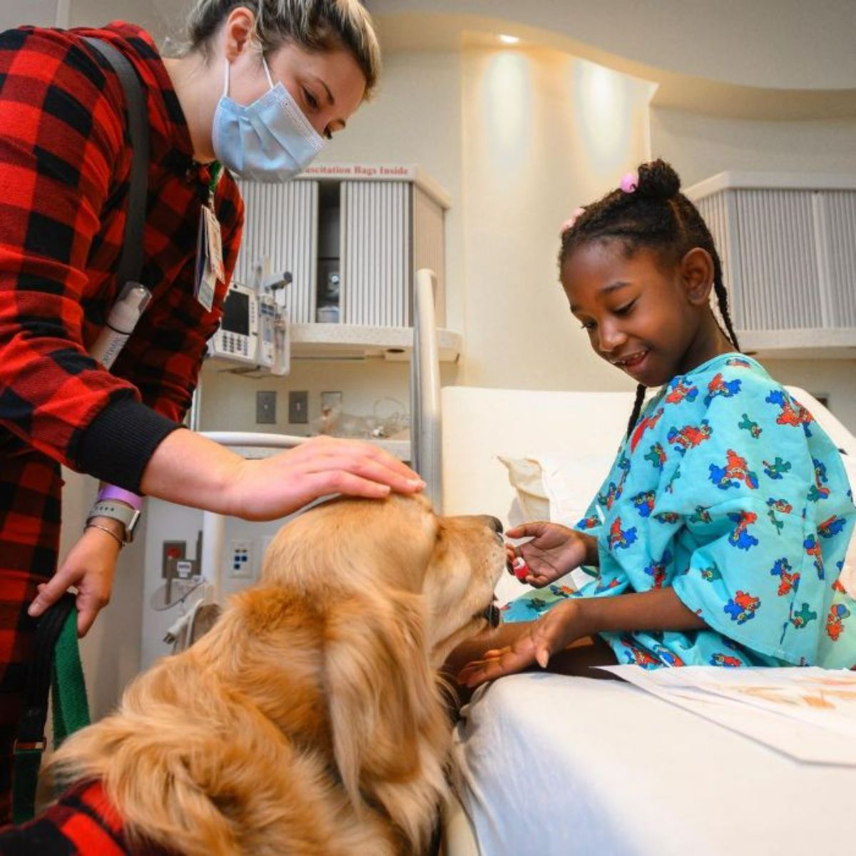 little african american girl sitting on a bed and touching a golden retriever sitting on the floor with a woman petting its head