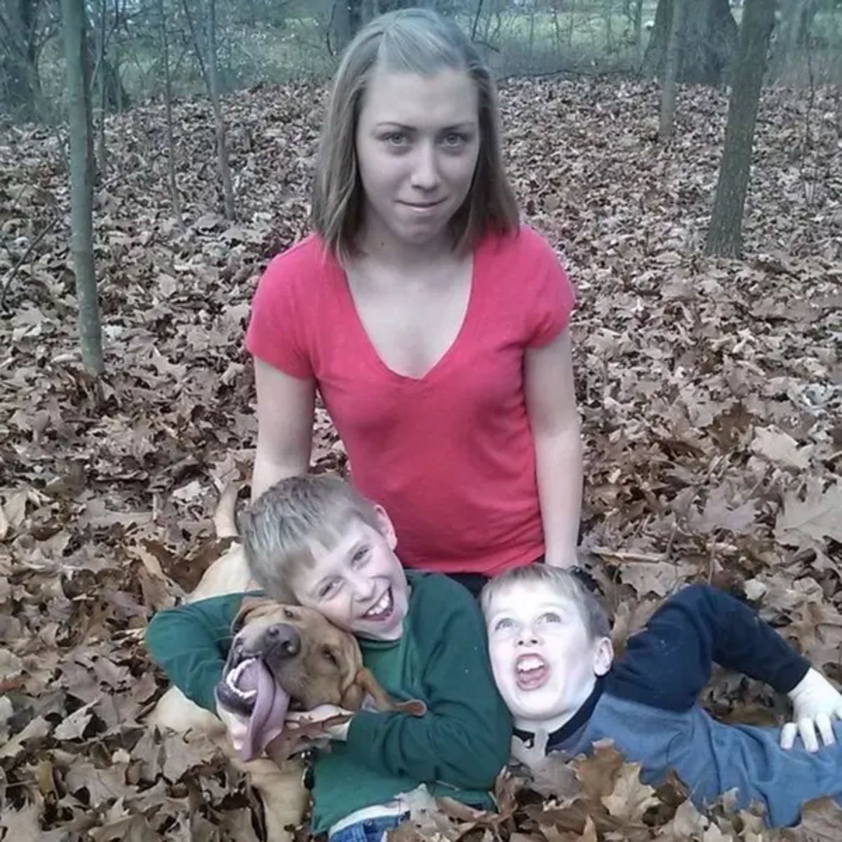 a woman sitting in a forest with brown leaves covering the ground with 2 boys and 1 boy holding a brown dog with the tongue hanging out