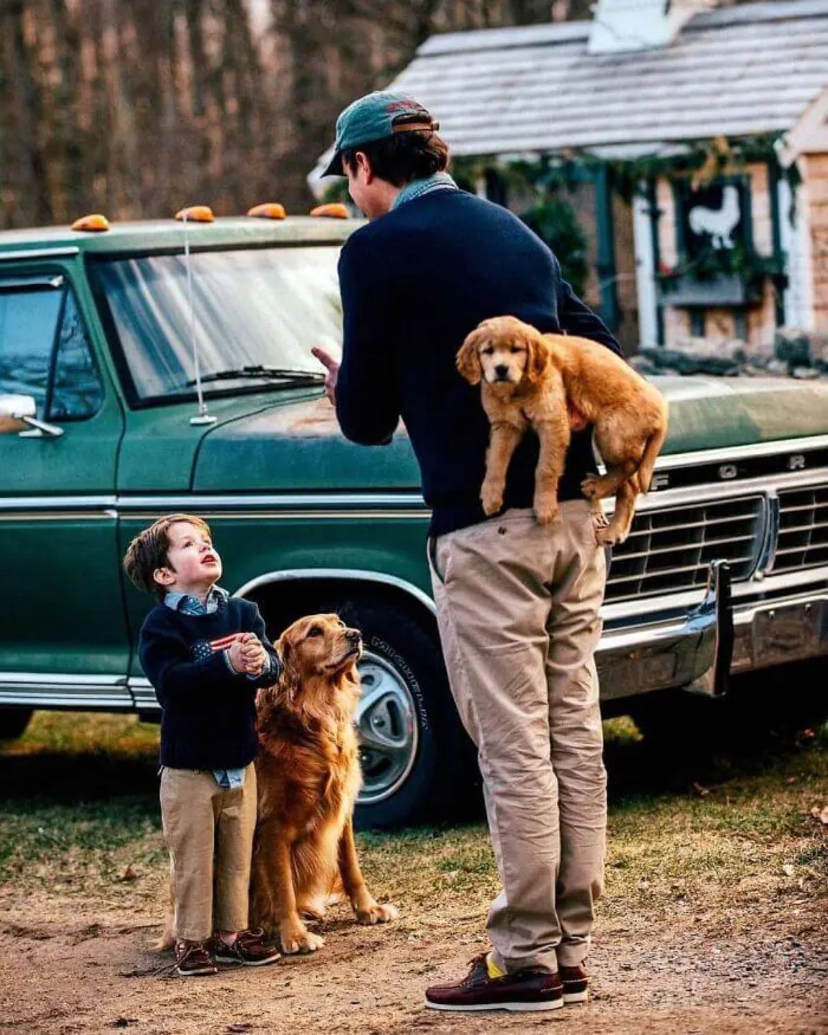 little boy standing next to a golden retriever sitting on grass in front of a man who is holding a golden retriever puppy behind his back