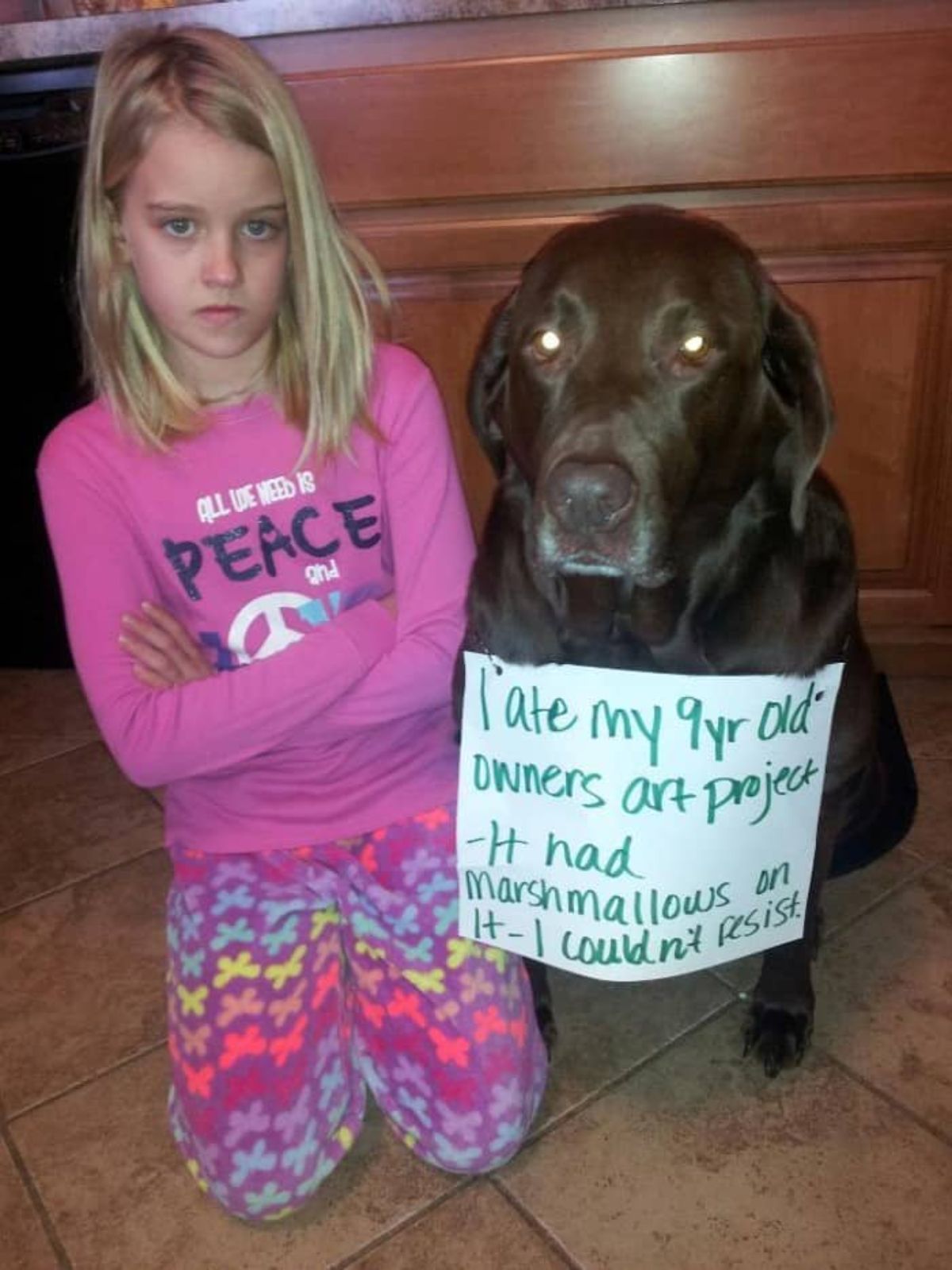 angry little girl kneeling by a black dog with a note around the neck saying I ate my 9 yr old owners art project- it had marshmallows on it- I couldn't resist