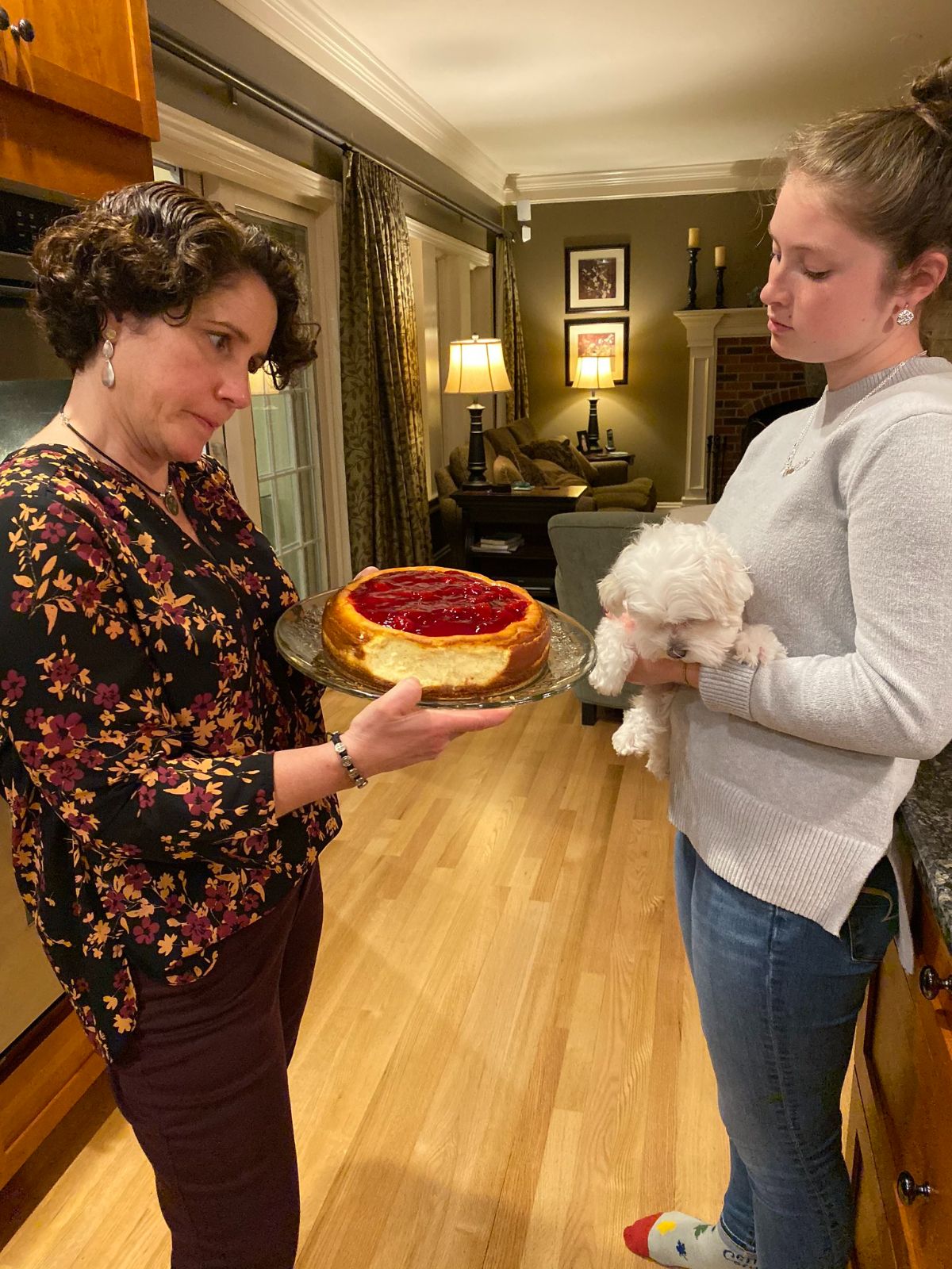 1 woman holding a small fluffy dog and another woman holding a cheesecake with one side eaten