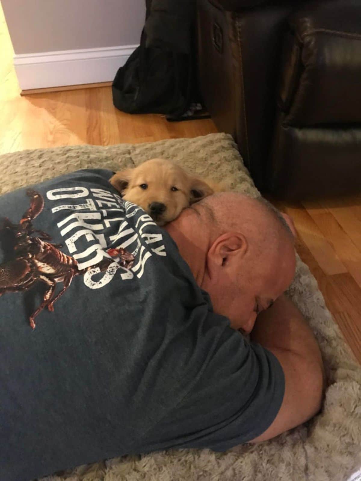 man laying on floor belly down hugging a golden retriever puppy who is peeking out from one side of the man's neck