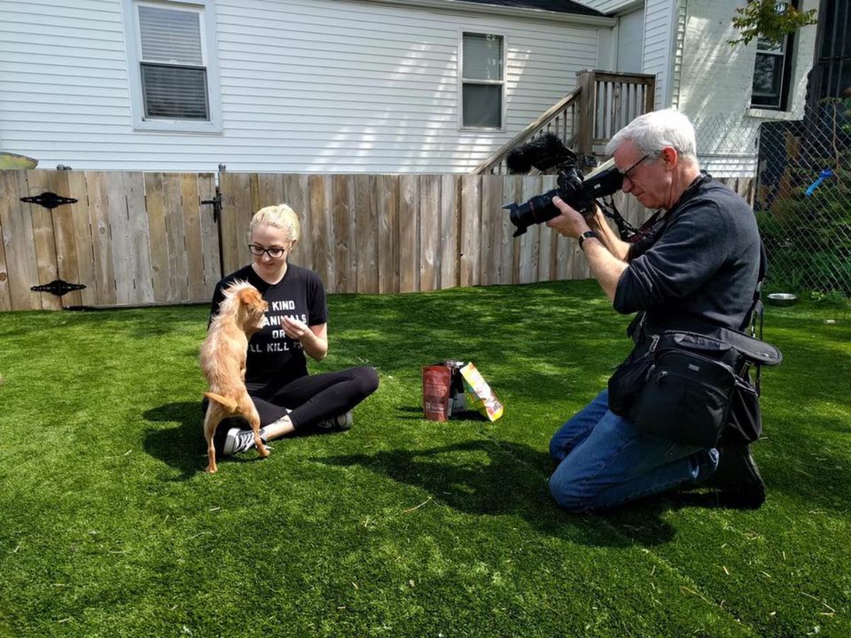woman sitting cross legged on grass with a small brown dog on hind legs while another person takes a photo with a camera
